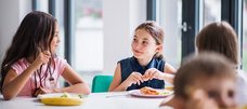 Zwei etwa zehnjährige Mädchen sitzen in der Schulmensa am Tisch und essen gemeinsam. Im Vordergrund sieht man noch andere Kinder. 