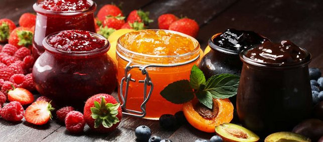 Verschiedene Marmeladen in Gläsern stehen auf einem Tisch und daneben liegen die entsprechenden frischen Früchte