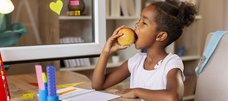 Schülerin sitzt zuhause am Schreibtisch und beisst genussvoll in einen Apfel