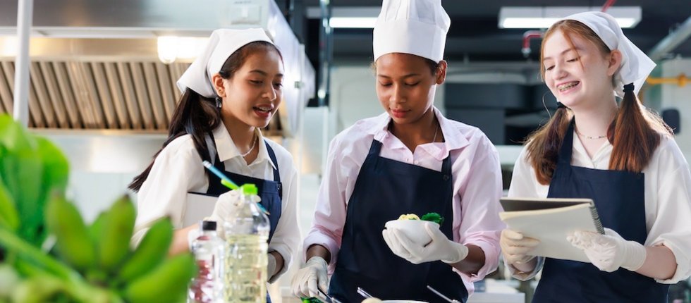 Drei Schülerinnen mit Kuchschürzen und Kochmützen kochen