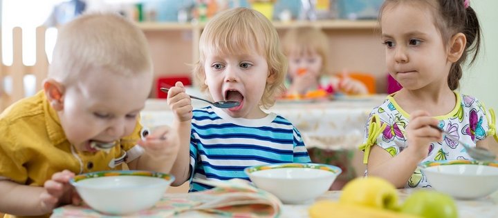 Drei etwa drei- bis vierjährige Kinder sitzen im Kindergarten zusammen an einem Tisch und essen mit Löffeln Suppe aus Suppenschalen. Im Vordergrund steht ein Teller mit einer Banane und zwei Äpfeln. 