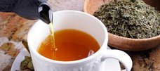 Tee fließt aus Kanne in Tasse, getrocknete Teeblätter daneben