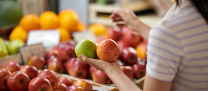 An einem Marktstand hält eine Frau zwei unterschiedliche Äpfel prüfend in der Hand. 