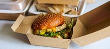 In einer Pappschachtel liegt ein vegetarischer Burger mit Salat und Käse. Im Hintergrund sieht man mehr Schachteln für Essen to-go. 