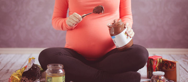 Schwangere Frau sitzt im Schneidersitz auf dem Boden, umgeben von Schokolade, sonstigem Süßkram und sauren Gurken, in der Hand ein großes Glas Schokocreme, aus dem sie gerade einen großen Löffel mit Creme herausgenommen hat.
