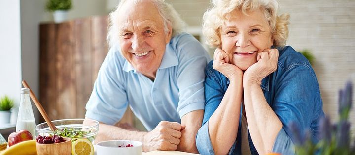 Eine Seniorin und ein Senior sitzen lächeln an einem Tisch, auf dem gesunde Lebensmittel liegen.