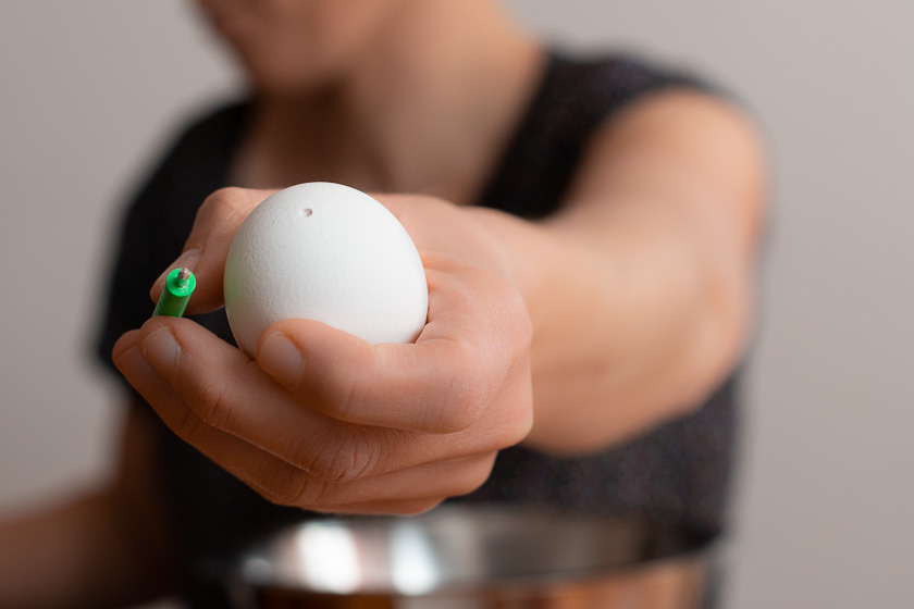 Eine Hand hält das Ausblasset – einen Miniblasebalg mit dünner Kanüle – ins Bild, die andere ein ausgeblasenes weißes Ei mit winzigem, sauberen Loch.