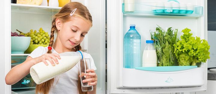 Mädchen steht vor geöffnetem Kühlschrank und giesst Milch ins Glas