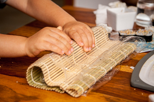 Kinderhände rollen das befüllte Nori-Blatt mit Hilfe einer Bambusmatte.