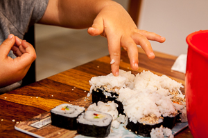 Kreatives Kochen: Kinderhände garnieren fertige Sushi-Stücke mit Bergen aus Sesam und Reis.