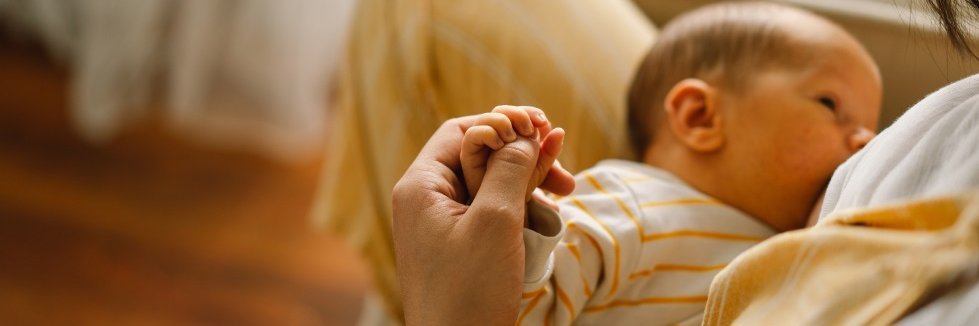 Ein Säugling trinkt an der Brust seiner Mutter und umklammert ihren Daumen mit seiner Hand.