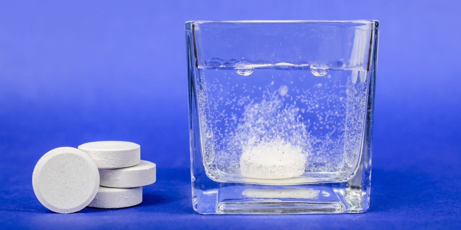 Vor einem blauen Hintergrund steht auf einem Tisch ein eckiges Trinkglas mit Wasser in dem sich eine Brausetablette sprudelnd auflöst. Neben dem Glas liegen weitere Brausetabletten.. Artikel "Überraschend viel Salz in Brausetabletten" öffnen.