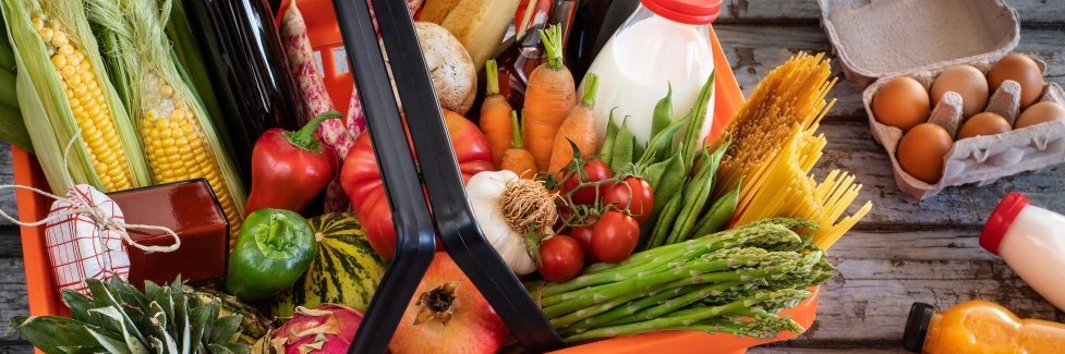 In einem roten Supermarktkorb liegen Paprika, Möhren, Mais, Tomaten, Spargel, Bohnen, Knoblauch und Spaghetti, ein Baguette, Milch, Öl, Ananas und ein Granatapfel. Daneben liegen Eier in einer Schachtel und Getränkeflaschen.