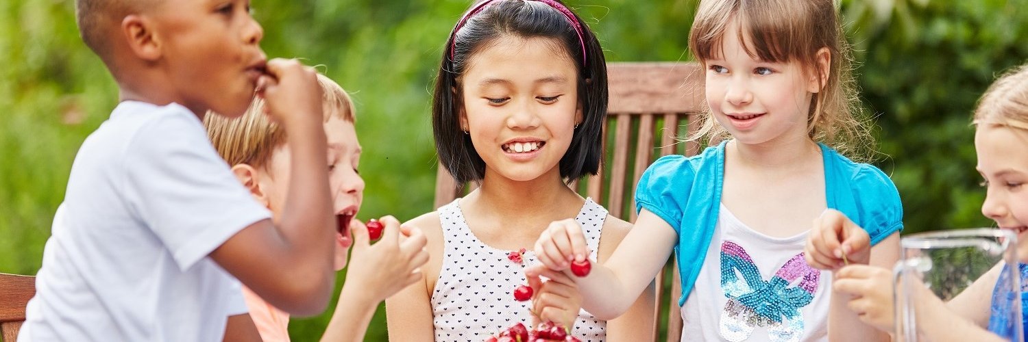 Fünf Kinder im Grundschulalter sitzen draußen gemeinsam an einem Tisch und essen Kirschen. 