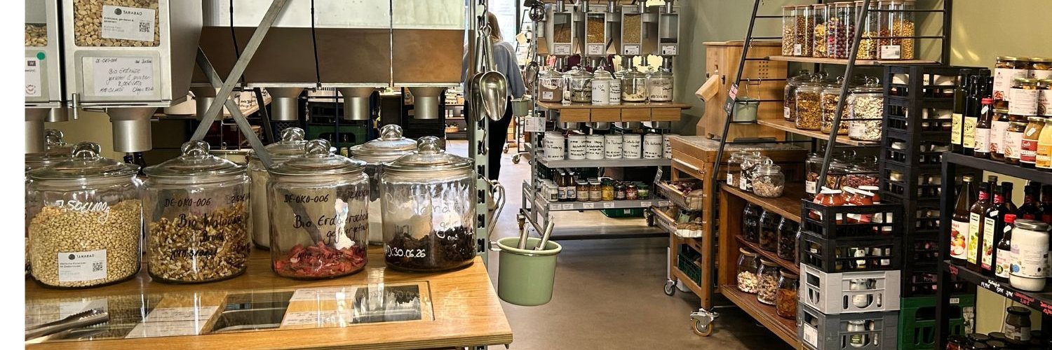 In einem Geschäft sind viele lose Lebensmittel in Gläsern oder durchsichtigen Behältern gelagert, die auf Holzregalen und Tischen stehen. Manches wird auch in Spendern angeboten oder abgefüllt in Gläsern und Flaschen.