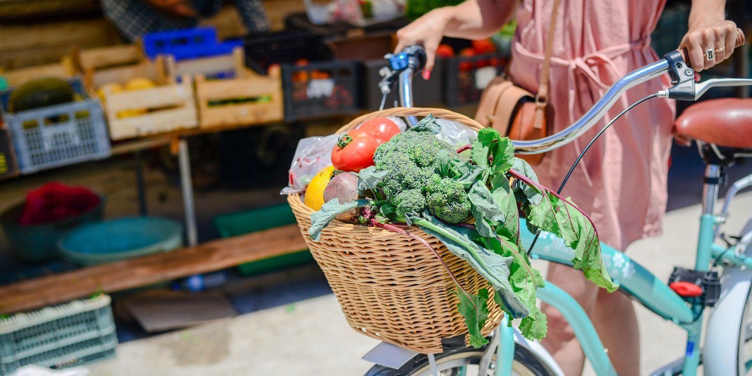 Eine Frau schiebt ein Fahrrad, an dessen Lenkrad ein Korb mit Gemüse hängt.. Artikel "Nachhaltig und günstig einkaufen" öffnen.