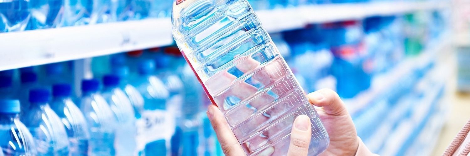 Vor einem Getränkeregal mit Wasserflaschen hält eine Frau eine PET-Flasche mit Mineralwasser in den Händen.