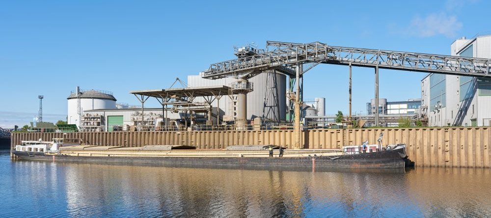 Ein langes Transportschiff liegt in einem Hafen und ein langes Rohr wird von oben in die geöffnete Ladefläche geführt.