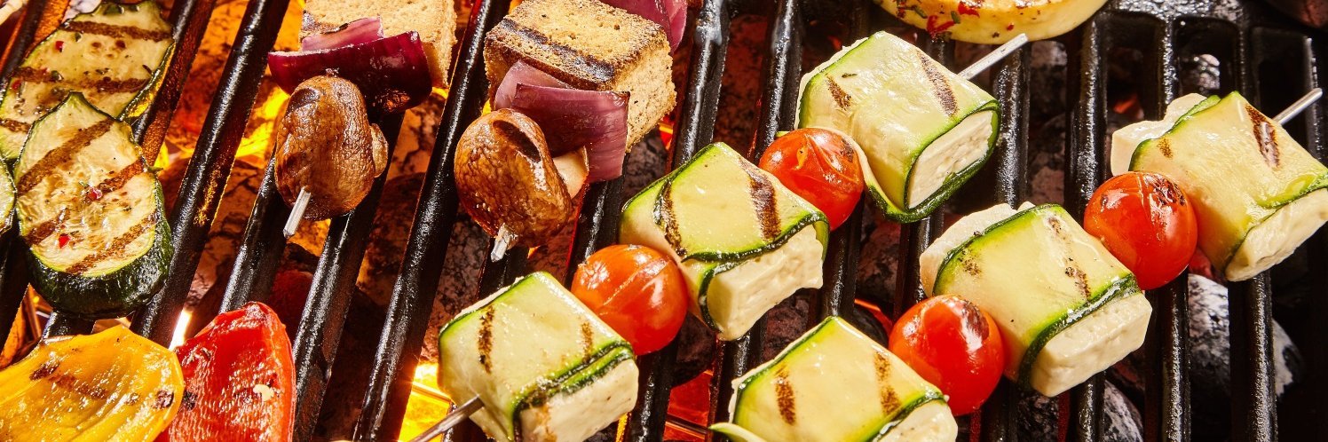Auf einem Grillrost liegen verschiedene Spieße mit Zwiebeln, Zucchinistreifen, Fetawürfeln und Cocktailtomaten sowie einzelne Paprikastücke.  