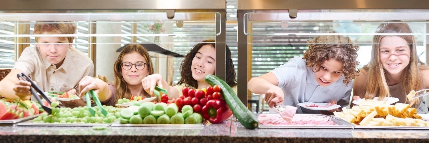 Drei Mädchen und zwei Jungen im Teenageralter bedienen sich an einer Essensausgabe mit frischem Obst und Gemüse.