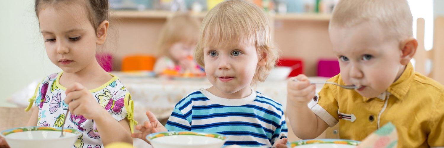 Drei Kleinkinder sitzen in einer Kita beim Essen am Tisch. Vor ihnen stehen tiefe Teller und eine Schale mit Äpfeln und Bananen.