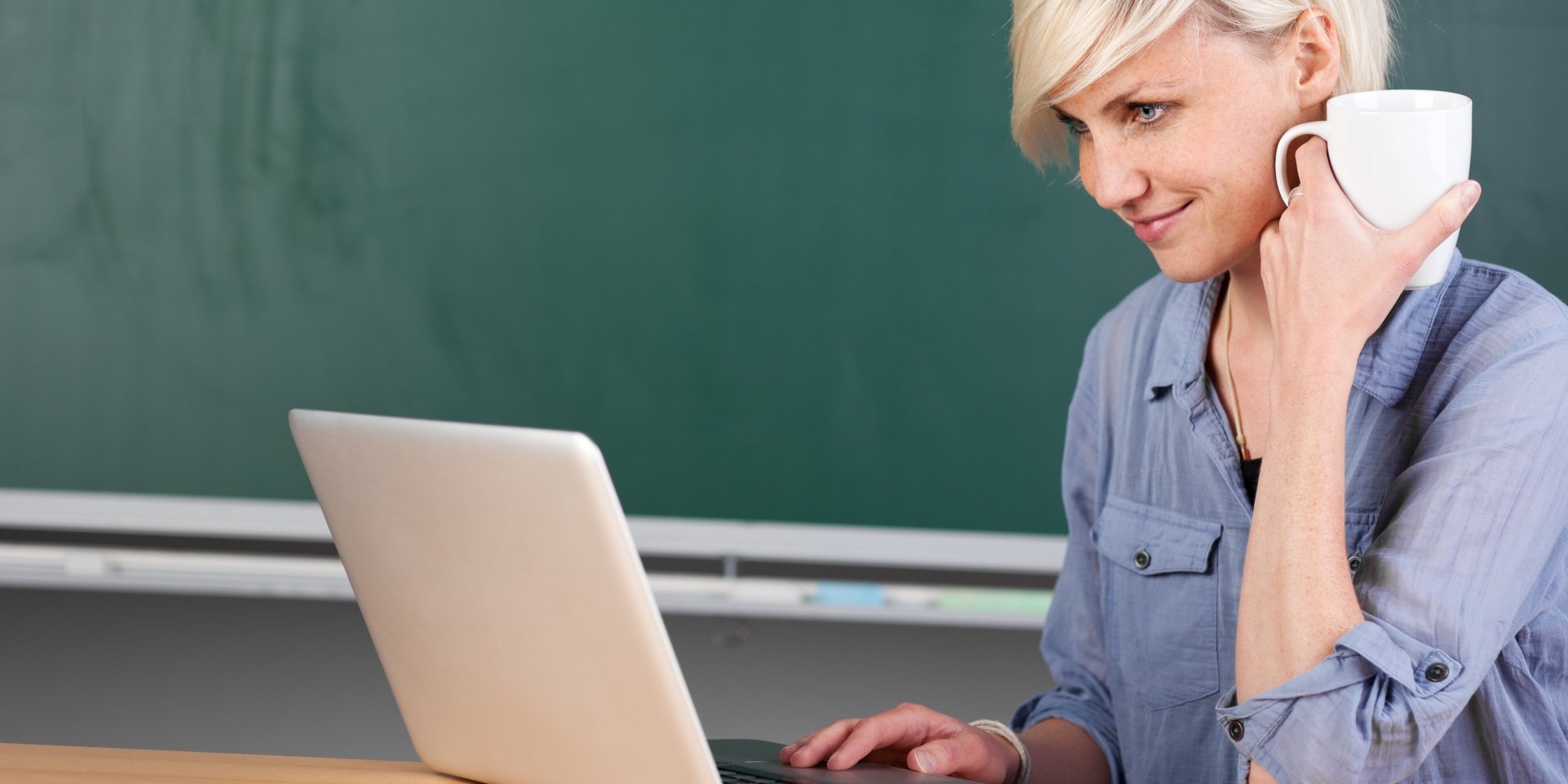 Eine Lehrerin sitzt am Tisch vor Tafel mit Laptop und Kaffeetasse.. Artikel "Fortbildungen" öffnen.
