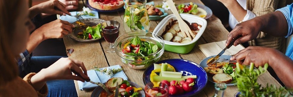 An einem großen Holztisch sitzen mindestens sechs Personen und essen und trinken gemeinsam. Auf dem Tisch stehen bunte vegetarische Gerichte. 