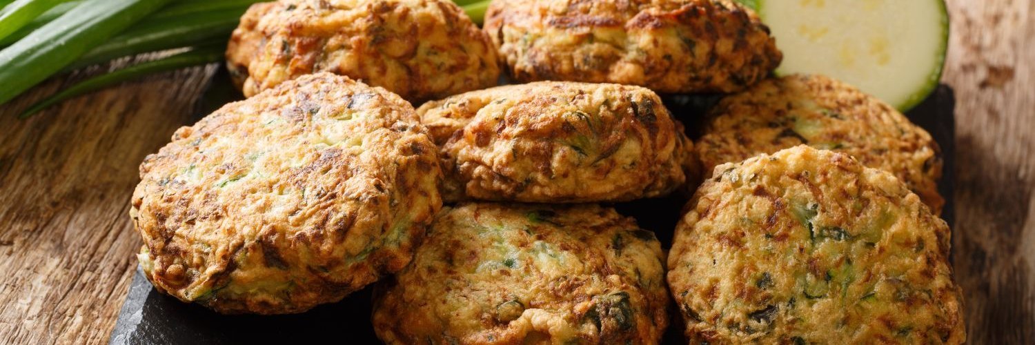 Auf einer schwarzen Schieferplatte liegen fertige Zucchinifrikadellen, daneben eine angeschnittene Zucchini und Frühlingslauch.
