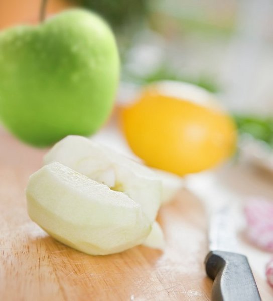 Auf einem Brett liegen ein halber geschälter Apfel, ein grüner Apfel, eine Zitrone und ein Küchenmesser.