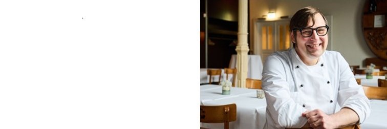 Ein Mann mit einer Kochjacke sitzt im Restaurant und laechelt in die Kamera.