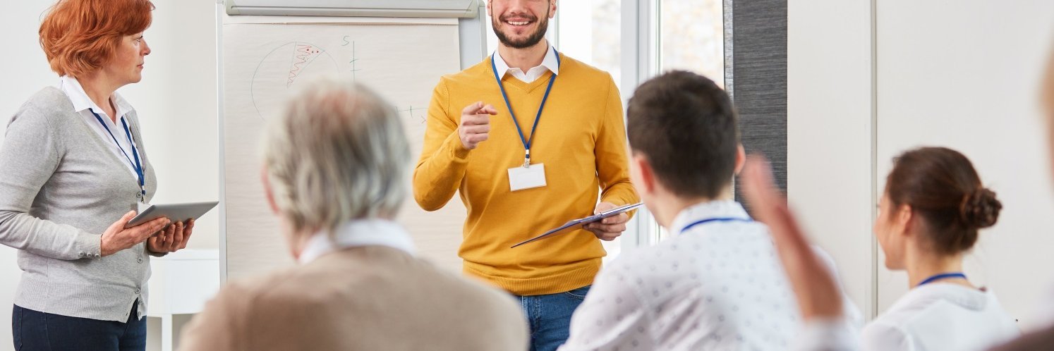 Ein Dozent im gelben Pullover steht vor einer Gruppe von Teilnehmenden in einem Seminarraum. Am Bildrand steht eine Frau mit Laptop in der Hand. 