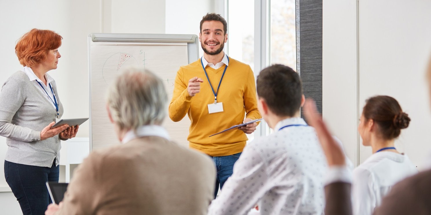 Ein Dozent im gelben Pullover steht vor einer Gruppe von Teilnehmenden in einem Seminarraum. Am Bildrand steht eine Frau mit Laptop in der Hand. . Artikel "Coaching für Kommunen und Schulen" öffnen.