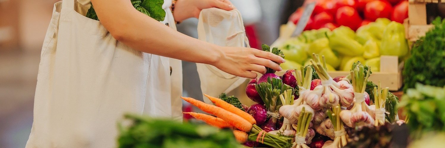 Eine Frau steht auf einem Markt vor einem Obst- und Gemüsestand und packt Gemüse in einen Leinenbeutel. 