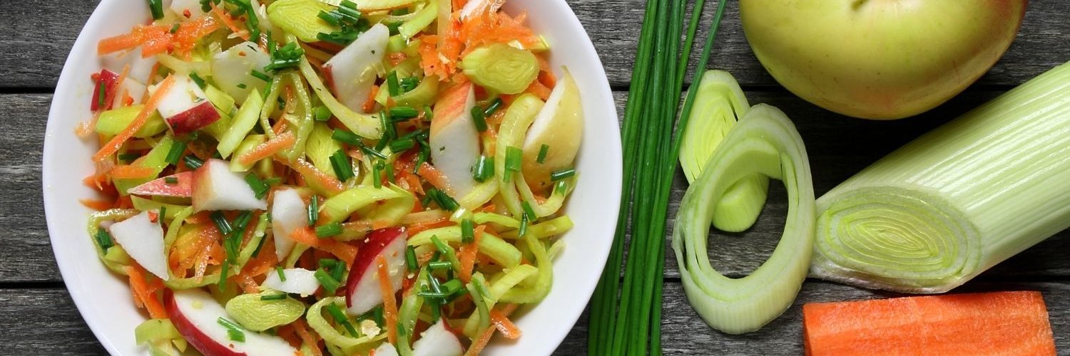 Auf einem dunklen Holzuntergrund steht eine weiße Schüssel mit Salat aus Lauch, Apfel, Möhre und Schnittlauch. Daneben liegen eine frische Lauchstange, ein Apfel, eine halbierte Möhre und Schnittlauch