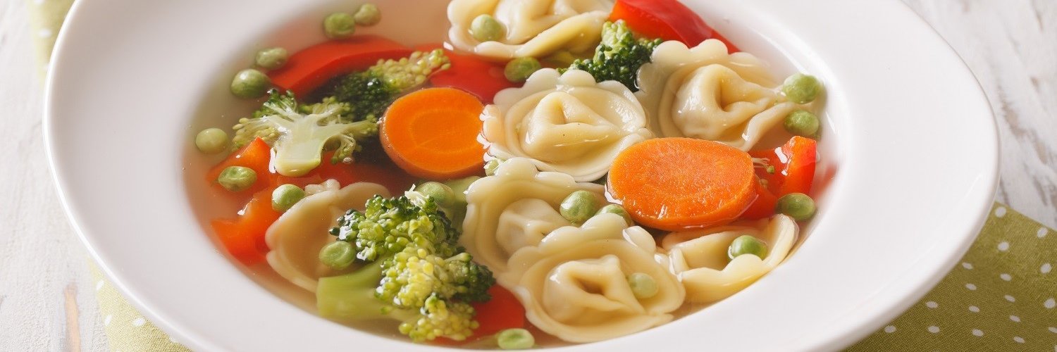 In einem weißen Suppenteller befindet sich eine Suppe mit Tortellini, Brokkoli, Erbsen, Karotten und Paprika. 