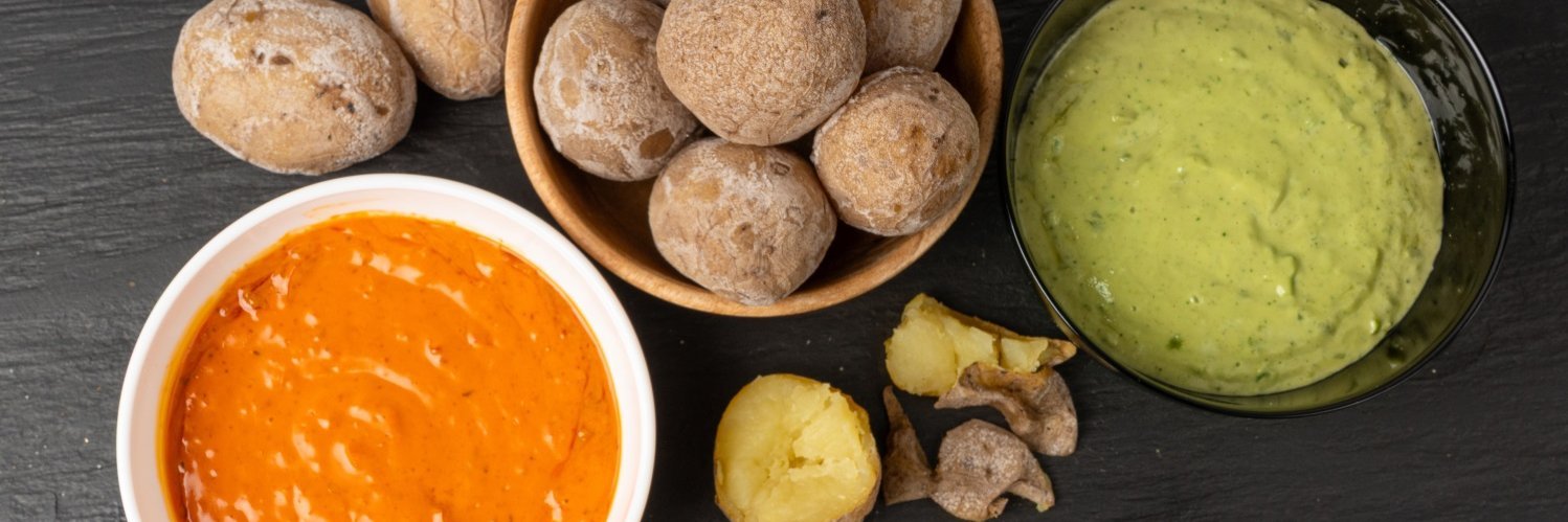 Auf einer schwarzen Schieferplatte steht eine Schale mit gebackenen Salzkartoffeln, eine halbierte Kartoffel liegt daneben. Links steht eine Schale mit rotem und rechts eine Schale mit grünem Dip.