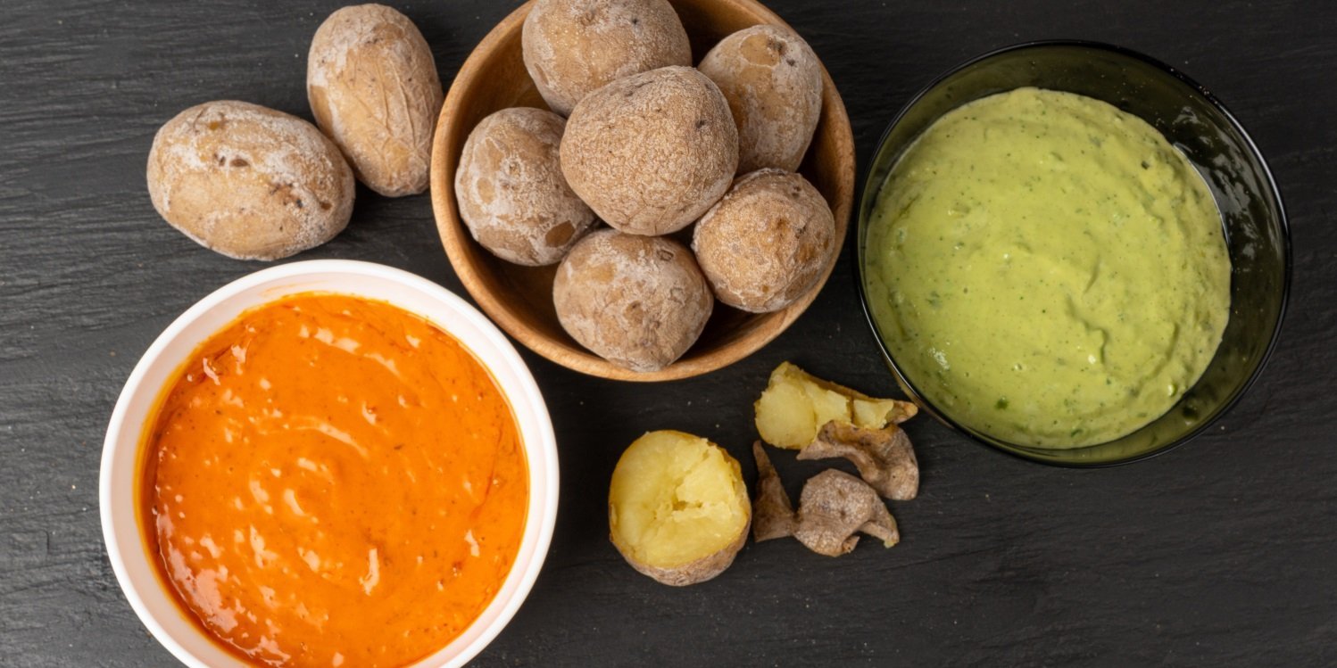 Auf einer schwarzen Schieferplatte steht eine Schale mit gebackenen Salzkartoffeln, eine halbierte Kartoffel liegt daneben. Links steht eine Schale mit rotem und rechts eine Schale mit grünem Dip.. Artikel "Kanarische Küche für zu Hause" öffnen.