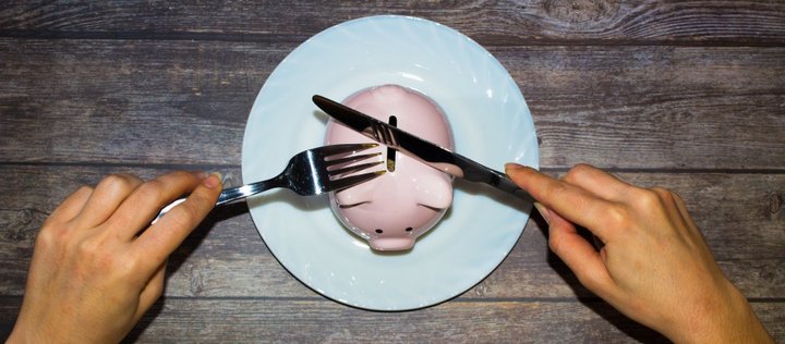 Auf einem Teller steht ein Sparschwein und eine Person versucht mit Messer und Gabel zu essen
