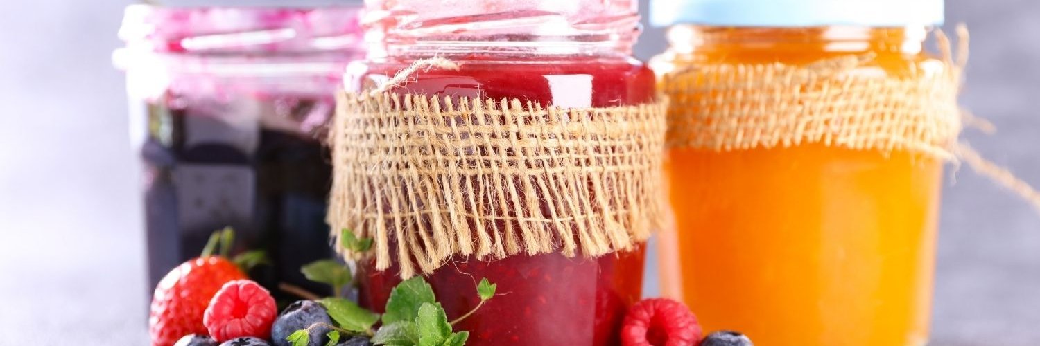 Vor einem grauen Hintergrund stehen drei Gläser mit Marmelade in Gelb, Rot und Lila, daneben liegen einige Beeren und Minzblätter.