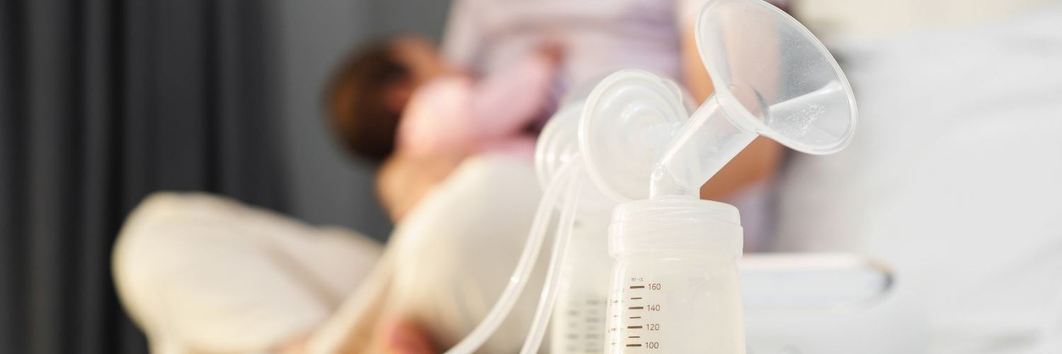 Auf einem Tisch steht eine Muttermilchpumpe mit Flasche und Inhalt. Im Hintergrund stillt eine Mutter ihr Baby.