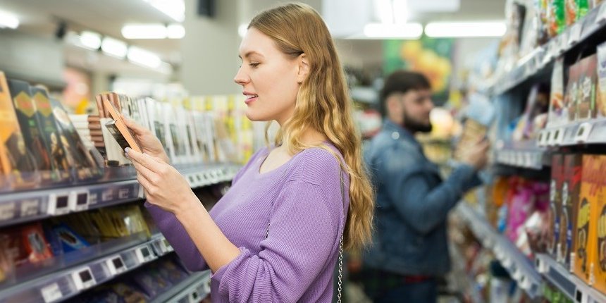 Eine junge Frau steht im Supermarkt vor dem Regal mit Schokolade. Sie hat eine Tafel in der Hand und liest das Etikett. . Artikel "Zucker bewusst genießen" öffnen.