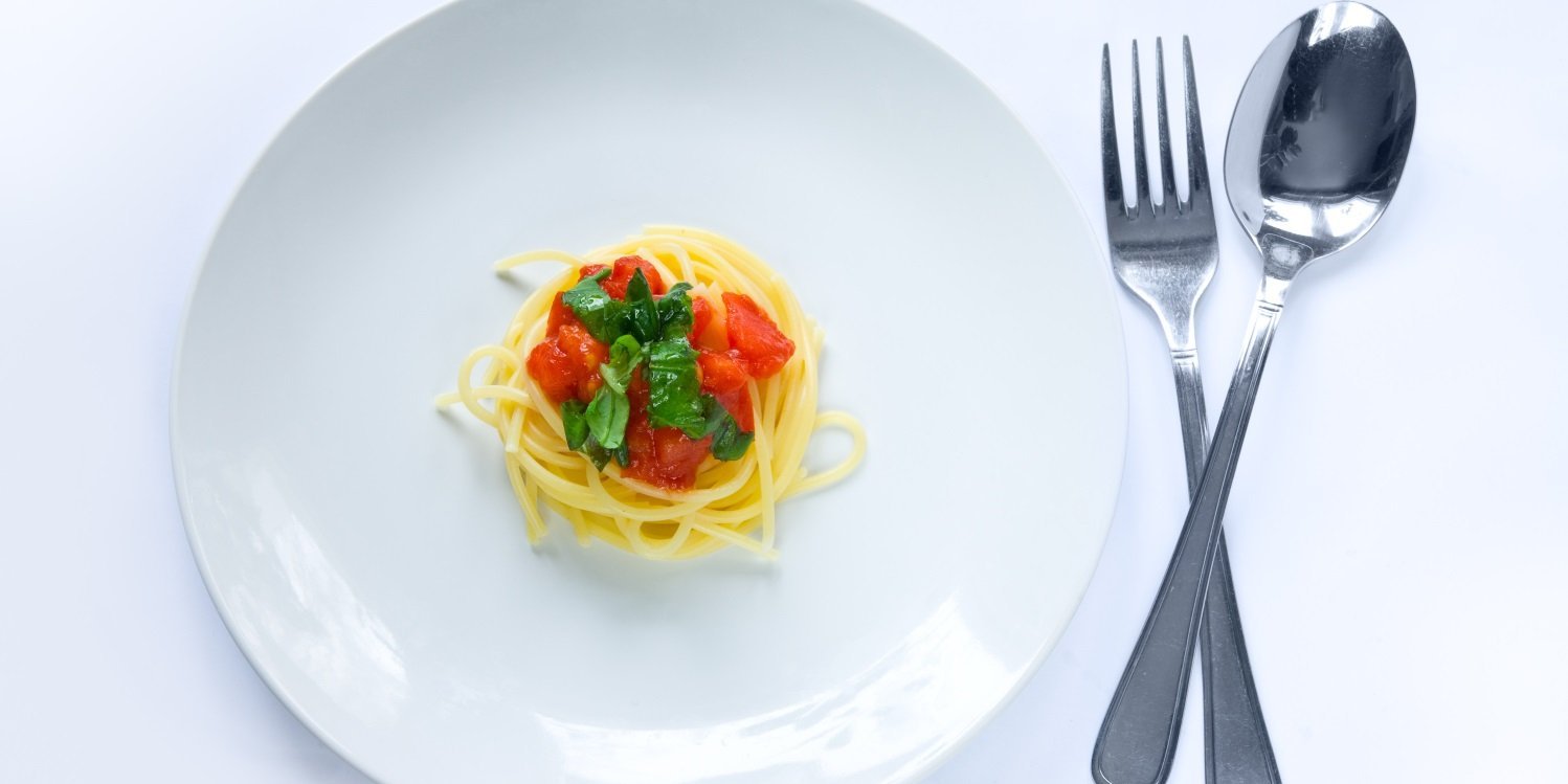 Auf einem weißen Teller liegt in der Mitte eine sehr kleine Portion Spaghetti mit einigen Tomatenwürfeln und Basilikum dekoriert, daneben Löffel und Gabel. . Artikel "Mini-Portionen im Trend?" öffnen.