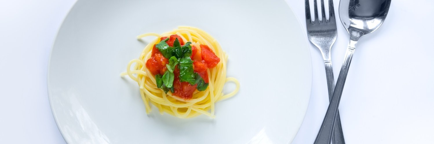 Auf einem weißen Teller liegt in der Mitte eine sehr kleine Portion Spaghetti mit einigen Tomatenwürfeln und Basilikum dekoriert, daneben Löffel und Gabel. 