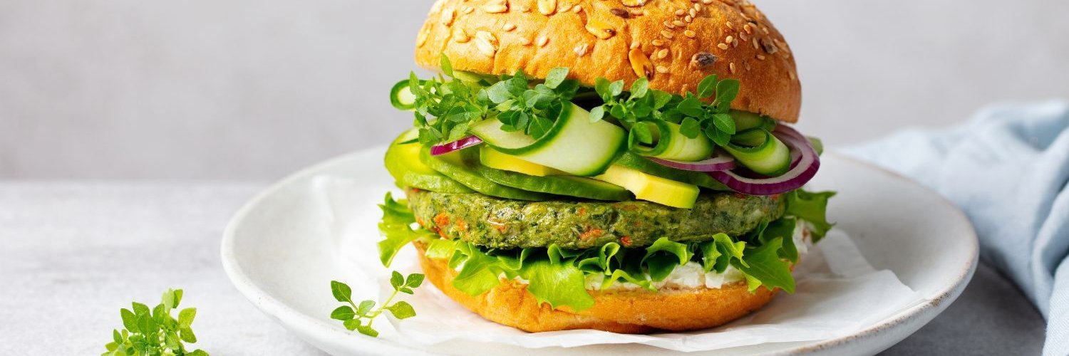 Auf einem weißen Teller liegt ein Burger mit einem Gemüsepatty, Gurken und Kräutern.
