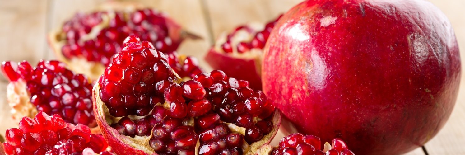 Auf einem hellen Holztisch liegt ein ganzer Granatapfel, daneben ein geöffneter in mehreren Stücken mit Samen darin.