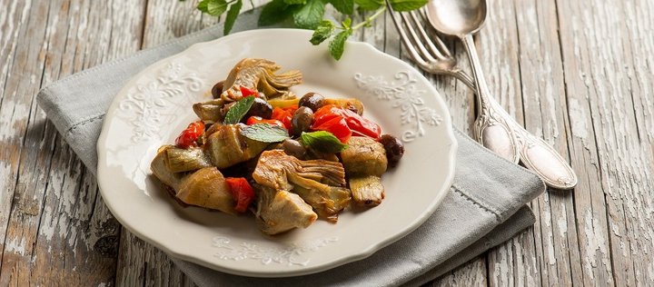 Auf einem weißen Teller auf einem Holztisch sind eingelegte Artischocken, Oliven und Tomaten angerichtet. 