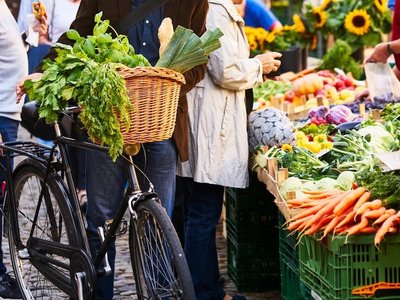 An einem Marktstand steht eine Person mit einem Fahrrad, an dem ein gefüllter Einkauskorb befestigt ist.
