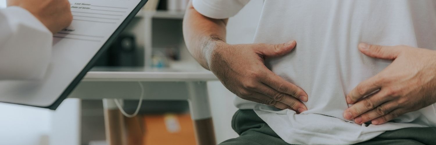 Ein Mann sitzt in einem Arztzimmer und hält sich mit beiden Händen den Unterbauch. Der Arzt neben ihm macht sich Notizen auf einem Klemmbrett.