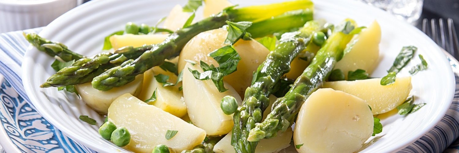Auf einem weißen Teller liegen gekochte Kartoffeln mit grünem Spargel, Erbsen und Kräutern. 