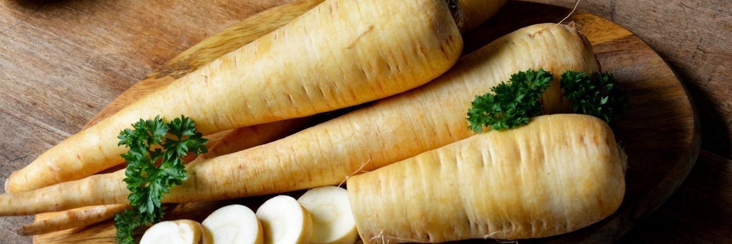 Auf einem Holzbrett liegen vier Pastinaken, eine davon ist zum Teil in Scheiben geschnitten.